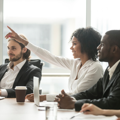 A woman with her raised hand in a business meeting with two men sitting on her left and right side. 
