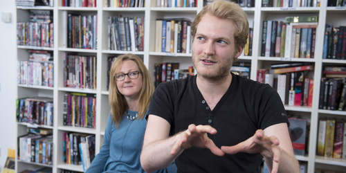 Man talking whilst using his hands for gesturing whilst a woman listening stands behind him in a library