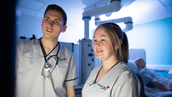 Two healthcare students in clinical uniforms review information on a monitor in a hospital training room, with medical equipment and a patient bed in the background