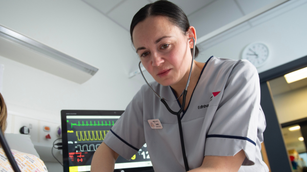 Student nurse using a stethoscope while monitoring a patient beside a vital signs monitor in a clinical setting.