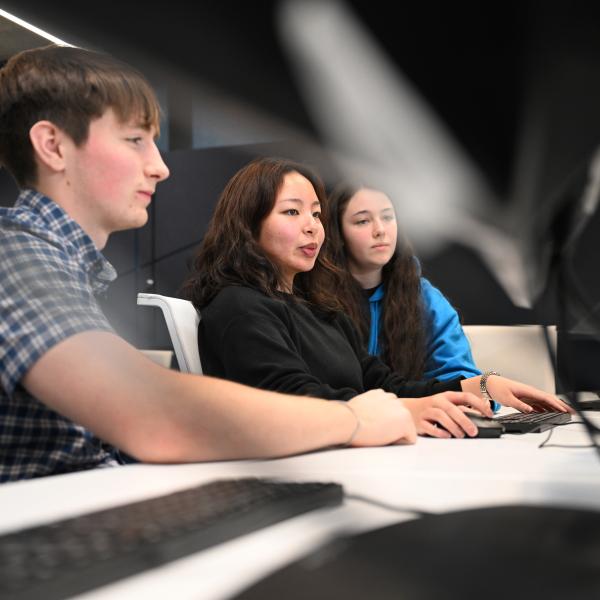 Three people sit side by side at a desk using desktop computers, focusing on screens in a modern indoor workspace. Keyboard and mouse are visible on the desk, with a blurred background and angled lighting overhead