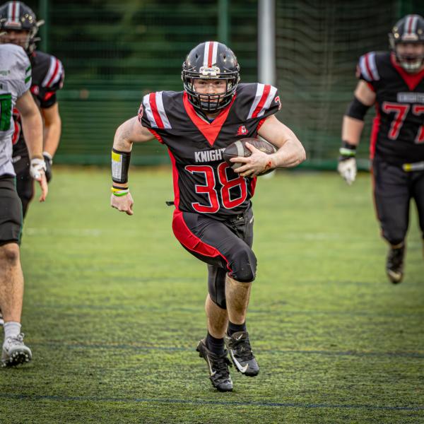 American football player wearing a black and red Napier kit with the number 36 runs forward holding the ball on a green field, while two other players in similar uniforms follow behind and an opposing player in a light-colored uniform approaches from the left.