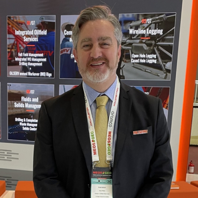 Gentleman in a dark business suit and tie wearing an exhibitor badge, standing at a trade show booth with oilfield services posters in the background.