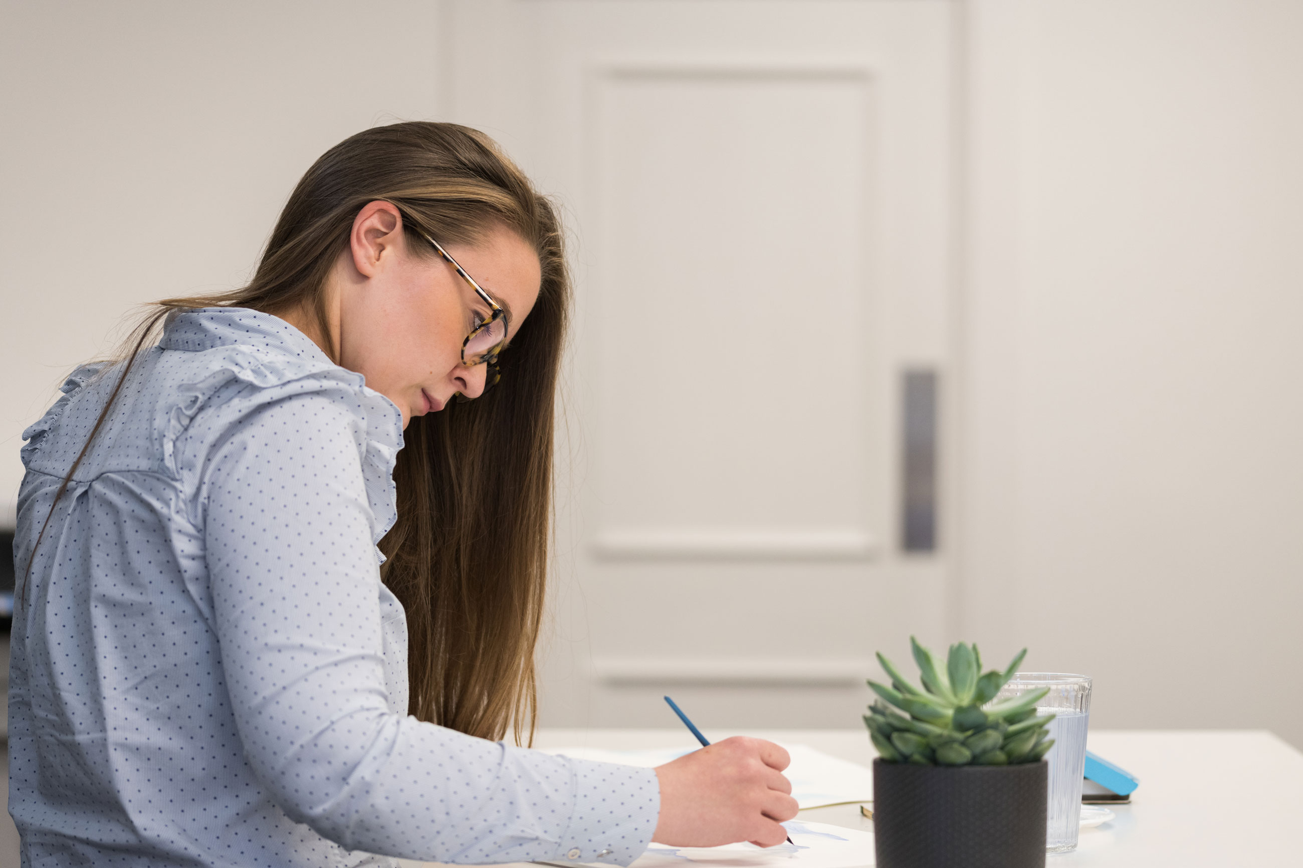 Melissa Preston sitting at a desk writing on a piece of paper. A green plant is placed on the table