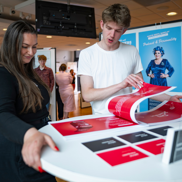 Two people reviewing printed materials at an indoor event table, with posters and displays in the background