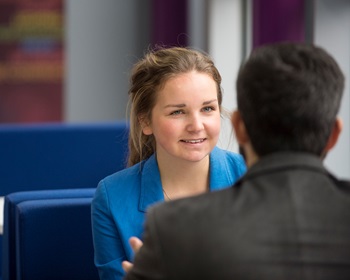 A women sitting on a sofa chatting with a male colleague. She is facing the camera, he has his back to the camera