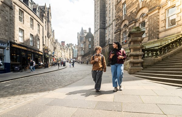 Two Edinburgh Napier students walking down the famous serpentine street in Edinburgh's old town city centre, smiling and talking in the sunlight. 