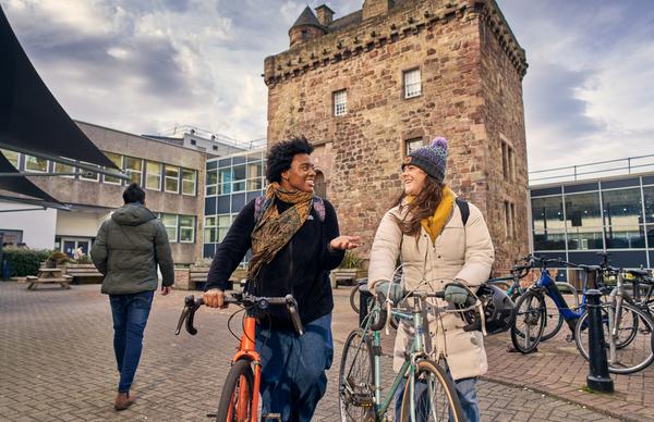 Two students talking while they leisurely push their bikes around at Edinburgh Napier University's Merchiston campus on a sunny evening. 