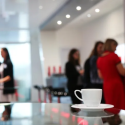 White coffee cup and saucer on a table in the foreground, with several people conversing in a softly blurred indoor meeting or networking space in the background
