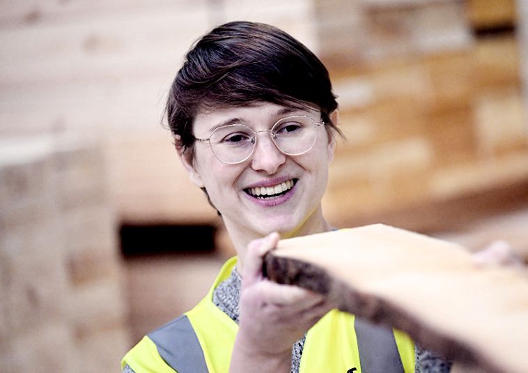 A person wearing a green high-visibility vest, smiling while holding a wooden plank.