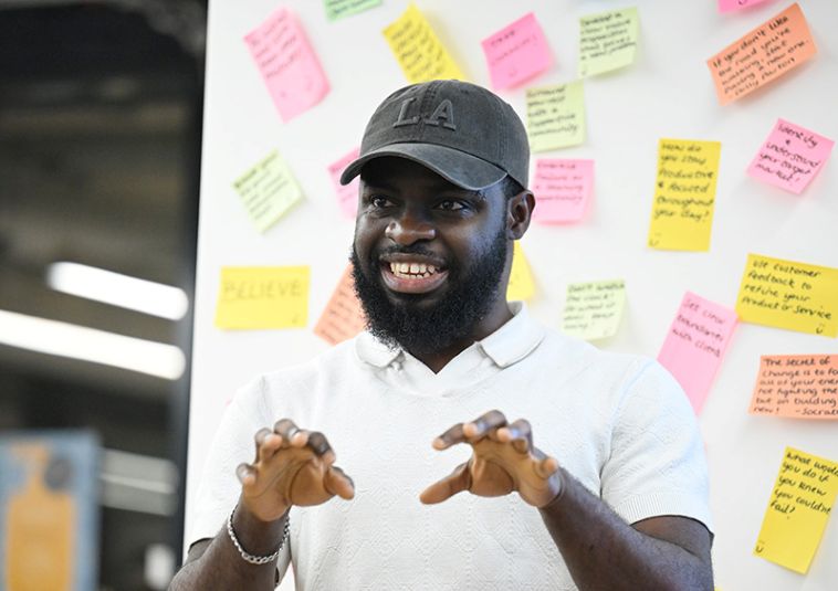 A person speaking and gesturing with their hands, with colourful Post-it notes in the background.