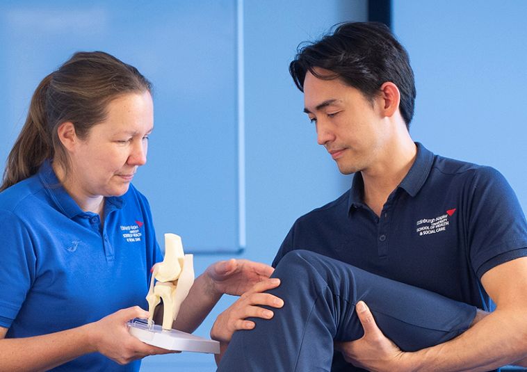 Two people dressed in shades of blue, with one person holding another person's leg while the other appears to provide care or assistance.