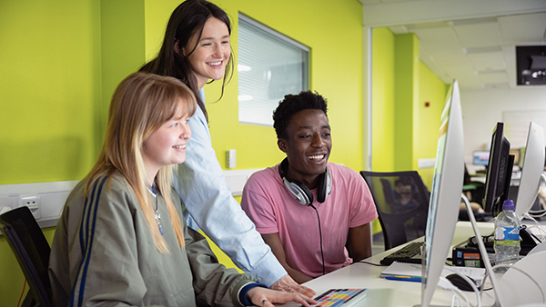 Three students are smiling at a computer screen whilst working together
