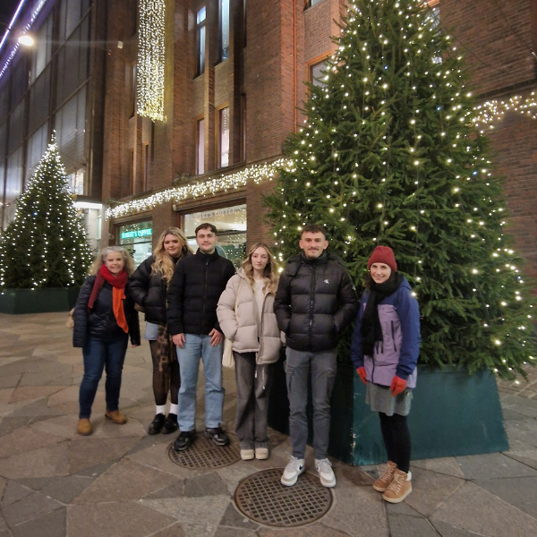 Group of six people standing outdoors beside large Christmas trees decorated with white lights in front of a brick building at night.