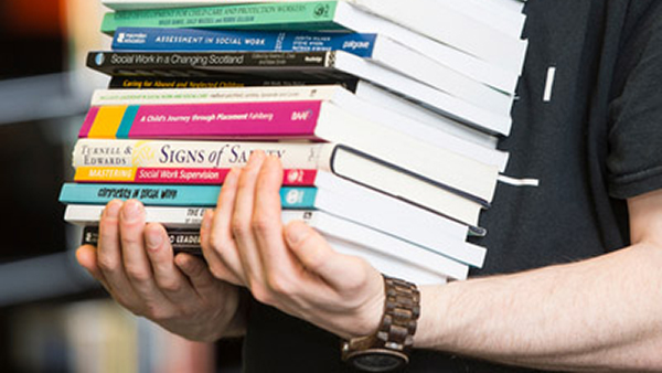 A student with just their arms visible underneath a huge pile of books